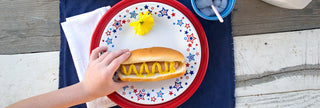 A hand holding a hot dog in a hot dog bun with only mustard on a red, white, blue star plate; on top of a scarlet plate with a blue placemat