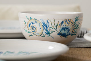 A Fiesta Retired Coastal Shores Bistro Coupe 5 1/2 Inch Cereal Bowl (22 OZ), decorated with painted seashell and seaweed designs in blue, yellow, and green, sits on a table set with plates and a gray napkin.