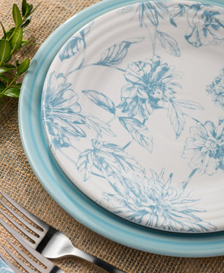 A close-up of two stacked plates with blue floral designs on a burlap placemat, next to a fork, knife, and a sprig of greenery.
