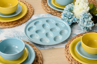 A pastel blue deviled egg tray sits on a floral-patterned tablecloth, surrounded by yellow and blue dishware on woven placemats, with a bouquet of blue and white flowers nearby.