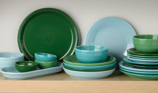A neatly stacked set of ceramic plates, bowls, and serving dishes in green and light blue hues sits on a wooden shelf against a light-colored wall.
