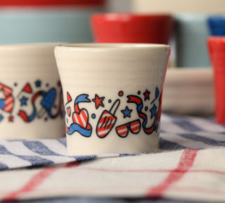 Three small white cups with red and blue star patterns are stacked in front of a pile of red, white, and blue plates and dishes, all arranged on a reflective surface.
