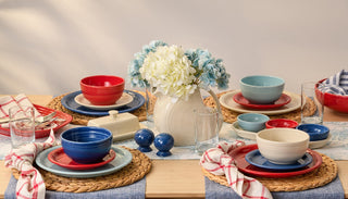 A dining table set with red, blue, and cream dishes, woven placemats, glasses, checked napkins, and a white pitcher filled with white and blue hydrangeas at the center.