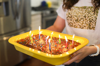 A person holds a yellow baking dish with a chocolate cake, colorful sprinkles, and lit candles in a kitchen. Only their torso and arms are visible.