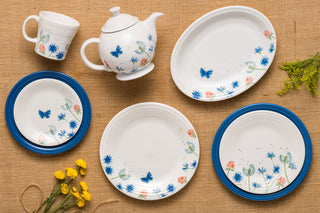 A set of white ceramic dishes, plates, a cup, and a teapot decorated with blue butterflies and floral patterns, arranged on a burlap surface with yellow flowers beside them.
