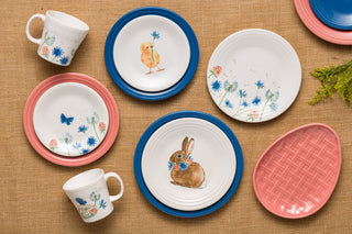 Colorful dinnerware set on a burlap surface, featuring plates and mugs with floral designs, a chick, a rabbit, and butterflies, plus a pink basket-weave patterned plate.