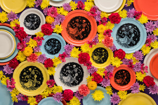 Colorful plates with Day of the Dead skeleton designs are arranged among vibrant yellow, pink, and purple flowers on a flat surface, creating a festive and lively display.