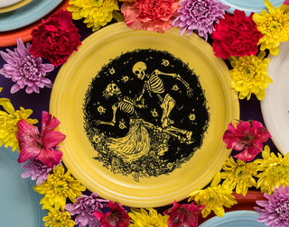 A yellow plate with a black illustration of two dancing skeletons surrounded by flowers, framed by colorful flowers and additional colorful plates around it.