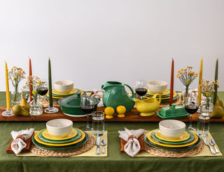 A colorful, elegant table setting with green, yellow, and cream dishes, wine glasses, candles, small vases of flowers, and napkins, all arranged neatly on a green tablecloth.