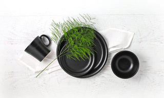 Black ceramic plates, bowl, and mug arranged on a white napkin and a white wooden surface, with a sprig of green foliage placed on top of the plates.