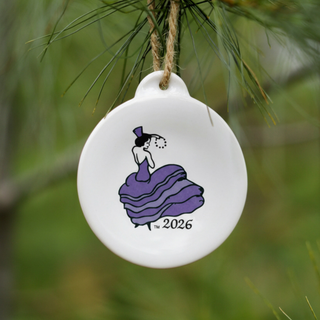 A round white ceramic ornament hanging from a pine branch, featuring a purple-dressed woman in a hat and the year 2026.