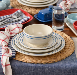 A place setting with stacked plates and a bowl with blue stripes, a glass of water, a napkin with red and blue plaid, on a woven placemat and blue tablecloth. Other tableware and dishes appear in the background.