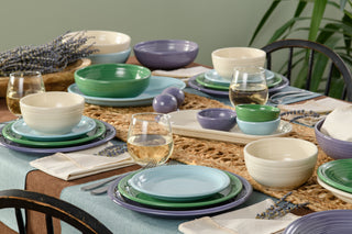 A dining table set with multicolored plates, bowls, and glasses of white wine, decorated with lavender sprigs and a woven placemat. The setting is cozy and inviting, with a plant in the background.