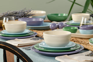 A dining table set with pastel plates, bowls, and glasses, decorated with lavender sprigs, woven placemats, napkins, and green plants in the background.
