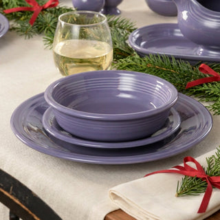 A purple bowl stacked on matching plates on a table decorated with evergreen sprigs, red ribbons, and a glass of white wine.