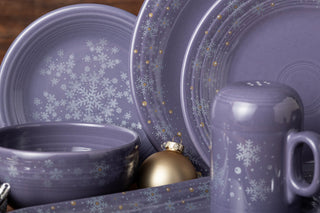 Purple ceramic dishes, a bowl, plate, and mug, all decorated with white snowflake patterns. A gold ornament is nestled among the dishes, emphasizing a winter or holiday theme.