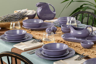 A dining table set with lavender-colored dishes, bowls, glasses of white wine, and a pitcher. Woven placemats, beige napkins, and a sprig of lavender decorate the setting.