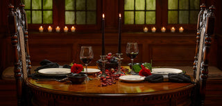 A romantic dinner table for two with black candles, rose petals, red roses, wine glasses, and plates, set in a dimly lit room with candles on the windowsill in the background.
