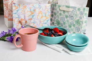 A pink mug of coffee, a turquoise bowl of assorted berries with a spoon, two stacked turquoise bowls, purple flowers, and floral gift bags on a white tablecloth.