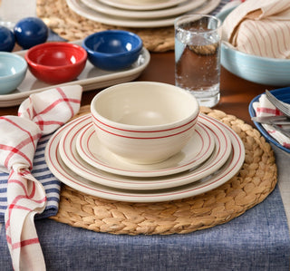 A table set with cream-colored dishes featuring red stripes, woven placemats, blue and red bowls, striped napkins, a glass of water, and bread in a blue dish, on a blue tablecloth.
