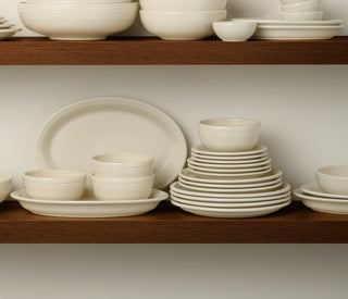 White ceramic bowls, plates, cups, and mugs are neatly stacked on a wooden shelf against a plain white background. The dishware is arranged in organized piles of different sizes.