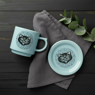 A light blue mug and matching saucer with black cat face designs are placed on a dark cloth napkin, next to green eucalyptus leaves, all set on a dark wooden surface.
