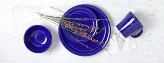 Bright blue ceramic plates, bowl, and mug arranged on a white napkin atop a white wooden surface, with a sprig of pink flowers placed across the plates.
