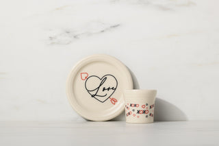 Small white plate with a heart and the word Love inside, next to a white cup decorated with red hearts and XOXO on a white marble surface and background.