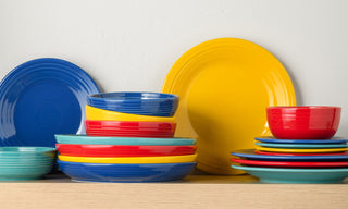 A collection of colorful plates and bowls in blue, yellow, red, and teal stacked on a wooden shelf against a white wall.