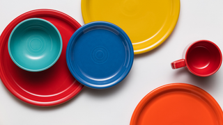 Colorful ceramic dishware, including plates, a bowl, and a mug, arranged on a white background. The dishes are red, blue, yellow, orange, and teal, overlapping in a vibrant composition.