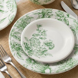 A green and white floral-patterned bowl and plates are stacked on a white napkin, set on a marble table with silverware, greenery, and a vase in a bright room near a window.
