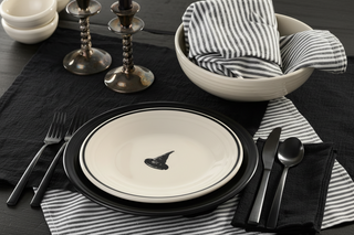 A table setting with black and white striped napkins, black cutlery, and a white plate featuring a witch hat graphic, all arranged on a black tablecloth.
