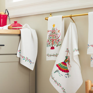 The Town and Country Holly Berry Christmas Tree Kitchen Towel Set features white cotton towels with festive embroidered designs, hanging on a gold towel bar by a light green cabinet topped with a red pot—perfect for holiday decor.
