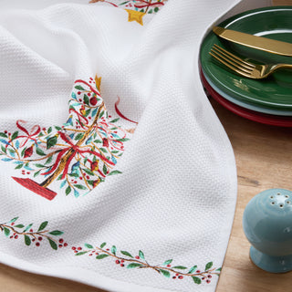 The Town and Country Holly Berry Christmas Tree Kitchen Towel Set, featuring a festive tree and berry design, is displayed on a wooden table beside green and red plates, gold utensils, and a blue salt shaker.