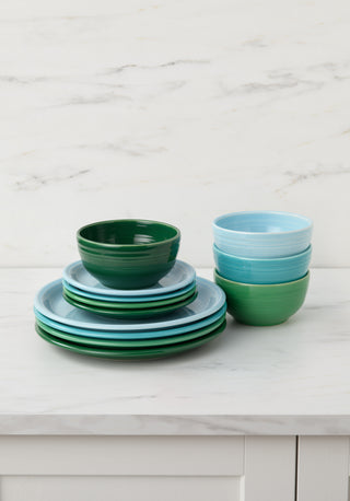 Stacked blue and green plates and bowls sit on a white kitchen counter against a white marble backsplash. The dinnerware is neatly arranged in two piles, one of plates and one of bowls.