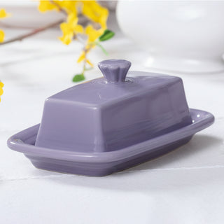 A Fiesta Lavender Extra Large Covered Butter Dish (8 1/8 x 5 1/8 in) sits on a white tablecloth, with yellow flowers and another white dish blurred in the background.