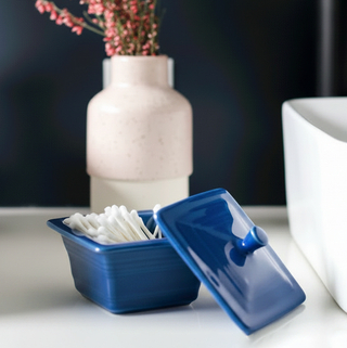 The Retired Fiesta Covered Box (4 5/8 Inch x 3 3/4 Inch) by Fiesta, in blue, holds cotton swabs on the bathroom counter beside a white vase filled with pink flowers and a white sink.