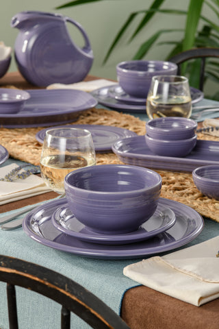 A dining table set with Fiesta’s Lavender Bistro Coupe 3-Piece Place Setting is arranged on a blue tablecloth, paired with woven placemats, cloth napkins, and two glasses of white wine.