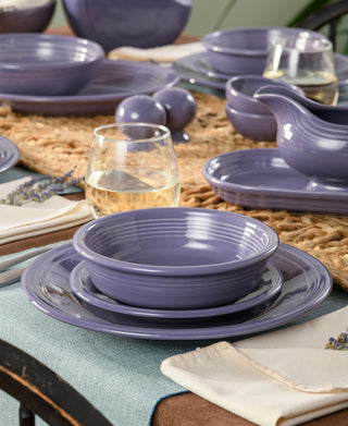A close-up of a dining table set with Fiesta's Lavender Classic Rim 3-Piece Place Setting, Service for 1, paired with glasses of white wine, white napkins, and a woven beige table runner.