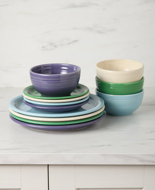 A stack of Fiesta’s Lavender Bloom Mixed Color Bistro Coupe 12-Piece Dinnerware, Service for 4, in purple, blue, green, and cream sits on a white marble countertop with a matching backsplash in the background.