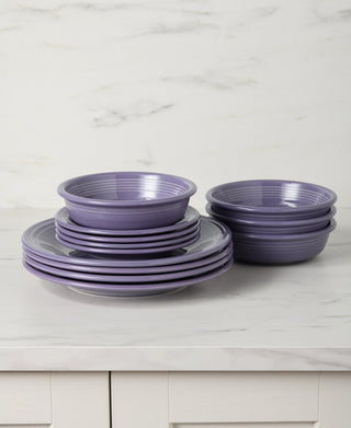 The Fiesta Lavender Classic Rim 12-Piece Dinnerware Set, Service for 4, featuring stacked plates and bowls, is arranged neatly on a white marble countertop with light cabinets below and a marble wall behind.