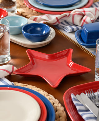 The Fiesta Star Plate, a red lead-safe china piece, is set among colorful dishes, glassware, and striped napkins. Blue, red, and cream plates combine with a woven placemat—perfect for sharing appetizers or gifting to a hostess.