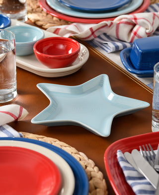 The Fiesta Star Plate, a light blue, lead-safe china piece, sits on a brown placemat amid red and blue dishes, water glasses, woven chargers, and striped napkins for a festive table setting.