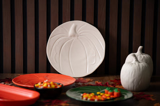 The Fiesta Pumpkin Shaped Plate 8 1/2 Inch in white stands upright against a dark striped background, surrounded by festive tableware, bowls of colorful candy, artificial autumn leaves, and a white pumpkin decoration.