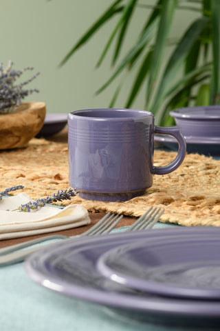 The Fiesta Lavender 16 OZ Stackable Mug rests on a woven placemat with coordinated plates, silverware, a white napkin, and lavender sprigs—ideal for a coffee bar setup accented by green plant leaves in the background.