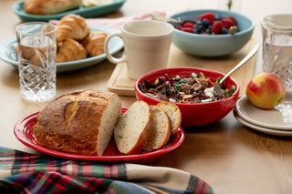A breakfast table features sliced bread on a red Fiesta 11 5/8" Oval Platter, salad with a fork in the Fiesta 38 oz Bistro Bowl, mixed berries, croissants, glasses of water, a mug, a peach, and a plaid cloth.