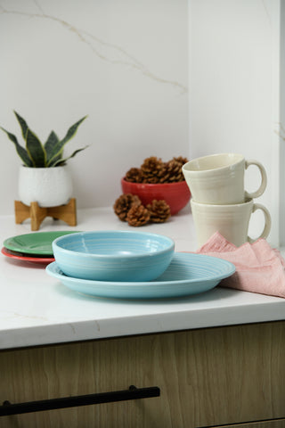 A stack of turquoise plates and bowls sits on a kitchen counter beside cream-colored mugs, a pink cloth, a potted plant, and a red bowl of pinecones. The countertop is white with a light wood cabinet below.