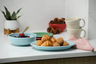 A breakfast scene features the Fiesta 2-Piece Serve Set, including an 11 5/8” oval platter and a 38 oz Bistro Bowl filled with mixed berries, alongside croissants, stacked mugs, a pink napkin, a plant, pinecones in a red bowl on a white countertop.
