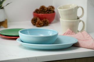 A 2-Piece Fiesta Serve Set—an 11 5/8 inch oval platter and a 38 oz bistro bowl—sits on a white countertop next to a pink napkin, with cream mugs, green plates, and pine cones in the background.