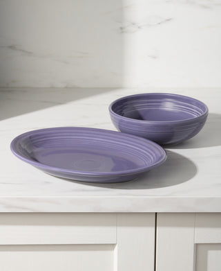 A Fiesta Lavender 2-Piece Serve Set—an 11 5/8 inch oval platter and a 38 oz Bistro bowl—rests on a white marble countertop with light streaming in from the left and soft shadows on a light-colored wall in the background.